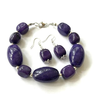 Puede incluir: Un conjunto de joyas con una pulsera y pendientes a juego. La pulsera y los pendientes están hechos de grandes cuentas ovaladas de color púrpura oscuro. La pulsera tiene un cierre plateado y los pendientes tienen ganchos plateados.