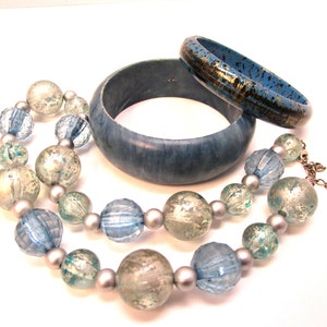 Puede incluir: Un collar de cuentas azules y plateadas con dos brazaletes de madera azul. Las cuentas son iridiscentes y tienen un aspecto moteado. Los brazaletes son lisos y tienen un efecto de mármol.