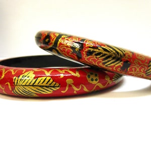 May include: Two red wooden bangles with black and gold painted designs. The bangles are stacked on top of each other.