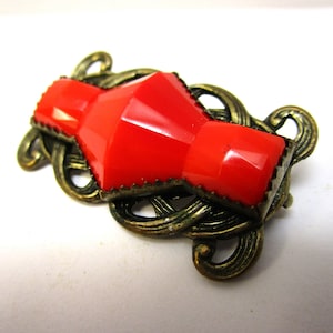 May include: A red glass brooch with a geometric design set in a gold-toned metal frame with intricate swirls.