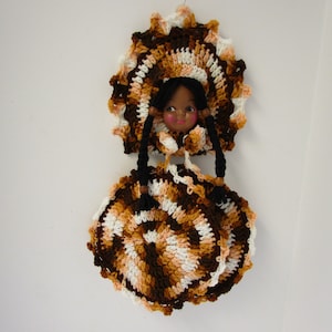May include: A handcrafted doll featuring a crocheted dress and bonnet. The doll's face is painted with rosy cheeks and a sweet expression. The dress and bonnet are in shades of brown, white, and peach.