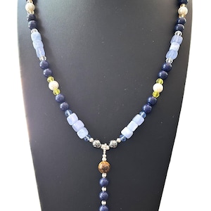 Puede incluir: Un collar de cuentas con una variedad de cuentas azules, blancas y amarillas. El collar tiene un colgante con una piedra marrón y beige y una hebra de cuentas azules. El collar se exhibe en un soporte negro.