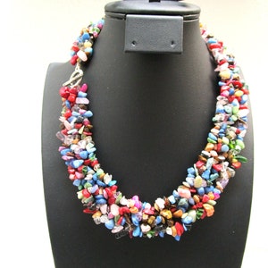 Peut inclure: Un collier multicolore composé de petites pierres de formes irrégulières. Les pierres, aux couleurs vives comme le rouge, le bleu, le vert et le jaune, sont présentées sur un présentoir noir, mettant en valeur leur aspect naturel.