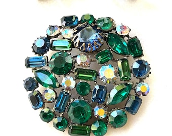 May include: Vintage jewelry set featuring a large brooch and matching clip-on earrings. The brooch is a circular design with an array of emerald green, sapphire blue, and iridescent crystals. The earrings have a similar design with green and blue crystals.