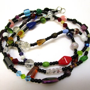 Puede incluir: Un collar de múltiples hilos con cuentas negras y cuentas de vidrio de colores. El collar tiene un cierre plateado.