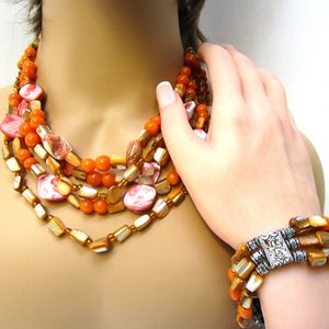 Peut inclure: Ensemble collier et bracelet multi-rangs composé de perles de coquillage orange et rose. Le collier est composé de plusieurs rangs de perles, dont des perles rondes orange et des perles de coquillage plates plus grandes avec des motifs roses et blancs. Le bracelet est de conception similaire, avec un fermoir argenté.