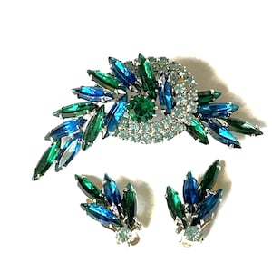 Könnte beinhalten: Ein Vintage-Broschen- und Ohrring-Set mit einem Wirbeldesign aus blauen und grünen Strasssteinen. Die Brosche ist größer und hat einen zentralen grünen Strassstein, der von kleineren blauen und klaren Strasssteinen umgeben ist. Die Ohrringe sind kleiner und haben ein ähnliches Design mit blauen und grünen Strasssteinen.