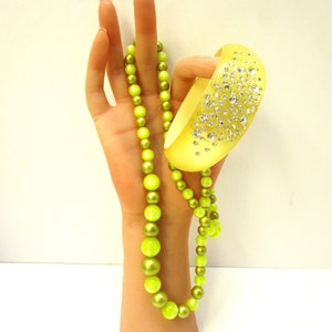 Puede incluir: Un collar de cuentas de color verde lima con una pulsera de brazalete de color verde lima con diamantes de imitación transparentes. La joyería se muestra en una mano.