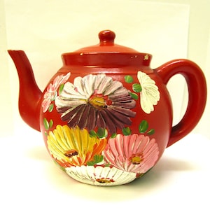 Könnte beinhalten: Eine rote Teekanne mit einem floralen Design, das auf die Seite gemalt ist. Die Blumen sind in den Farben Rosa, Weiß, Gelb und Lila.