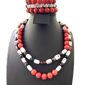 Puede incluir: Un conjunto de collar de dos hilos y pulseras a juego. El collar presenta cuentas rojas, blancas y negras. Las pulseras están apiladas encima del collar y tienen una combinación de colores similar. La joyería se exhibe en un soporte negro.