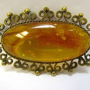Op de afbeelding: Een gouden filigraan broche met een grote ovale amber steen. De amber is diepgeel van kleur met rode en bruine insluitsels.