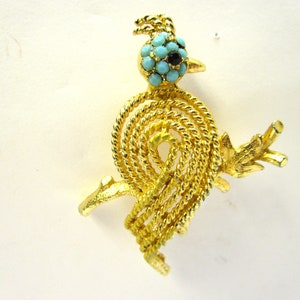May include: Gold-tone brooch featuring a stylized bird with a turquoise and black eye. The bird is depicted with a rope-like design and a twig-like branch.