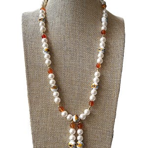 Puede incluir: Un collar largo de perlas blancas con cuentas de patrones naranja y blanco. El collar tiene un borla en la parte inferior con dos cuentas blancas y dos cuentas de patrones naranja y blanco.