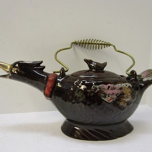 Può includere: Teiera in ceramica marrone a forma di anatra, con becco dorato e collare rosso. Ha un manico in ottone e un coperchio con un pomello a forma di uccello. Design vintage unico.