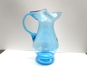 May include: A light blue glass pitcher with a curved handle and a ruffled rim. The pitcher has a round base and a small orange sticker. The pitcher is approximately 8 inches tall.