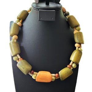 Puede incluir: Un collar artesanal con cuentas de madera verde oliva, naranja y natural. El collar se exhibe en un busto de joyería negro. Las cuentas son de varias formas y tamaños, creando una estética única y terrosa.