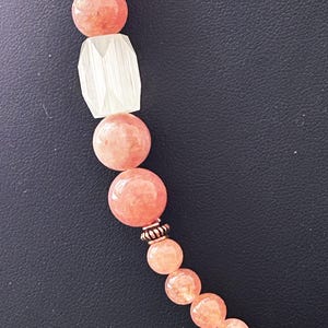 Puede incluir: Primer plano de un collar con grandes cuentas redondas de color melocotón intercaladas con una cuenta clara rectangular facetada. El collar tiene una cuenta espaciadora de color cobre y cuentas de melocotón más pequeñas.