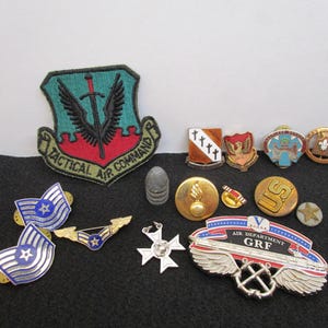 Puede incluir: Colección de insignias militares, incluyendo parches, pines y distintivos. Los artículos presentan diversos diseños, como alas, banderas y el texto "TACTICAL AIR COMMAND". Los colores incluyen rojo, azul, dorado y plateado.