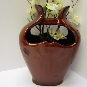 May include: A brown ceramic vase with a unique, abstract design. The vase has two large openings on the top and a smaller opening on the side. It is filled with white flowers.