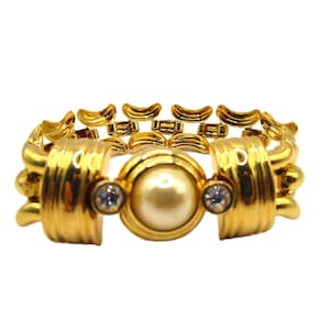 Op de afbeelding: Een gouden armband met een grote nepparel en twee heldere strass steentjes. De armband is gemaakt van met elkaar verbonden metalen stukken.