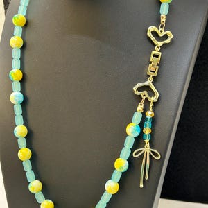 Puede incluir: Collar de cuentas con cuentas amarillas, azules y turquesas. Cuenta con un colgante dorado con dijes en forma de corazón y lazo. Las cuentas son redondas y rectangulares.