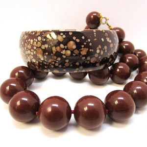 Puede incluir: Un brazalete de bangle moteado en marrón y blanco y un collar de cuentas marrones. Las cuentas son grandes y redondas.