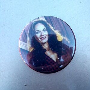 May include: A round button with a photo of a woman with long brown hair wearing a red and black plaid shirt. The woman is smiling and looking at the camera.