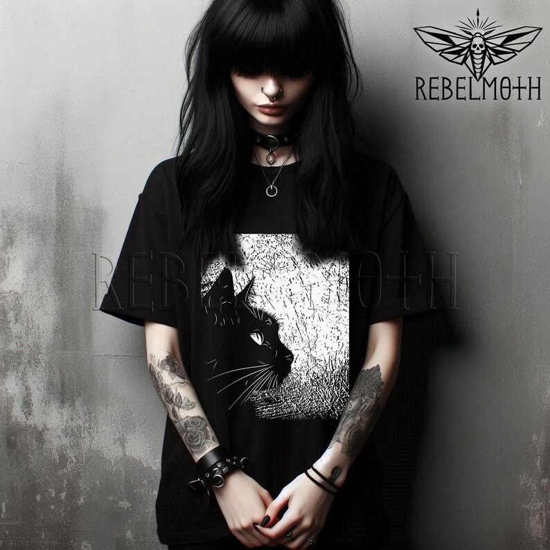 May include: A black short-sleeved t-shirt featuring a white graphic print of a cat profile against a textured background. The shirt displays the text REBELMOTH in the upper right corner, accompanied by a moth logo with a skull design.