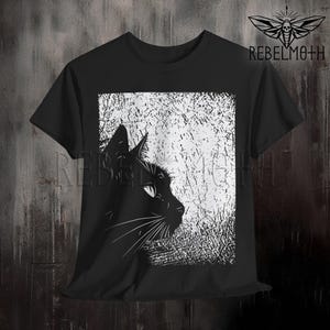 May include: A black short-sleeve t-shirt featuring a graphic print of a black cat in profile against a white, textured, rectangular background. The text REBELMOTH and a moth logo are printed in the upper right corner of the shirt design.