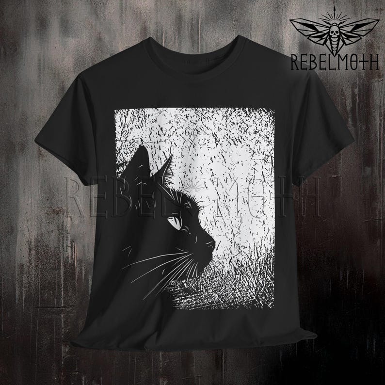 May include: A black short-sleeve t-shirt featuring a graphic print of a black cat in profile against a white, textured, rectangular background. The text REBELMOTH and a moth logo are printed in the upper right corner of the shirt design.