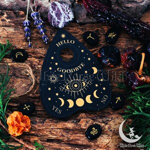 May include: Black wooden planchette with gold lettering and symbols, including "Hello", "Goodbye", "Yes", "No", and a crescent moon. The planchette is surrounded by black stones with various symbols, including a crescent moon, a star, and a cross.
