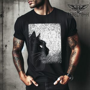 May include: A black short-sleeved t-shirt featuring a graphic print of a black cat profile against a white, textured rectangular background. The upper right corner displays a moth icon and the text REBELMOTH in a bold, black sans-serif font.