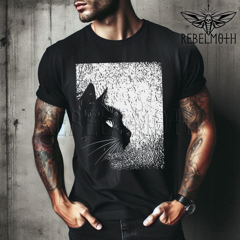 May include: A black short-sleeved t-shirt featuring a graphic print of a black cat profile against a white, textured rectangular background. The upper right corner displays a moth icon and the text REBELMOTH in a bold, black sans-serif font.