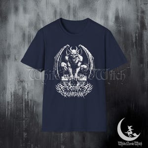 May include: Navy blue t-shirt featuring a white graphic of a gargoyle with spread wings, perched on a stone structure. Below the gargoyle is the text "DARK GOTHIC GUARDIAN" in a stylized font. The shirt has short sleeves.