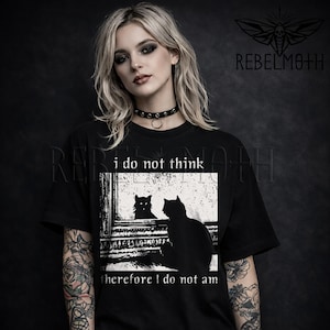 May include: A black short-sleeved t-shirt featuring a white graphic print of a cat looking into a mirror. The text above the graphic reads "i do not think" and the text below reads "therefore I do not am". The shirt has a crew neckline.