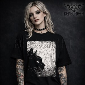 May include: A black t-shirt featuring a graphic print of a black cat in profile against a textured white background. The design includes the text REBELMOTH and a moth logo in the upper right corner. The shirt has a crew neckline and short sleeves.