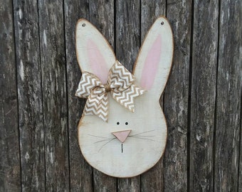 May include: A white wooden bunny rabbit head with pink ears and a brown and white bow. The bunny is painted with black eyes and a pink nose. The bunny is hanging on a wooden fence.
