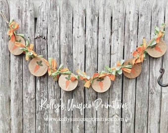 May include: A burlap banner with carrot designs and green and orange fabric accents. The banner is hanging on a weathered wooden fence. The text "Kelly's Unique Primitives" and "www.kellysunqiueprimitives.com" is printed on the fence.