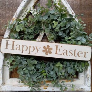 May include: A white wooden sign with the text "Happy Easter" and a small flower design. The sign is displayed in front of a green leafy wreath.