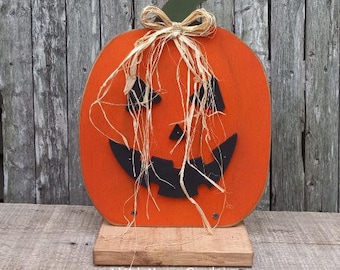 Könnte beinhalten: Ein hölzerner Kürbis mit einem schwarzen Jack-o'-Lantern-Gesicht und einer Strohbögen. Der Kürbis ist orange lackiert und hat einen grünen Stiel. Der Kürbis steht auf einem Holzsockel.