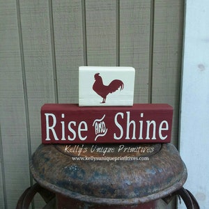May include: Red and white wooden sign with the text "Rise and Shine" and a rooster silhouette on a separate white block. The sign is sitting on top of a rusty metal container.