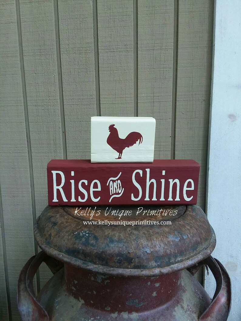 May include: Red and white wooden sign with the text "Rise and Shine" and a rooster silhouette on a separate white block. The sign is sitting on top of a rusty metal container.