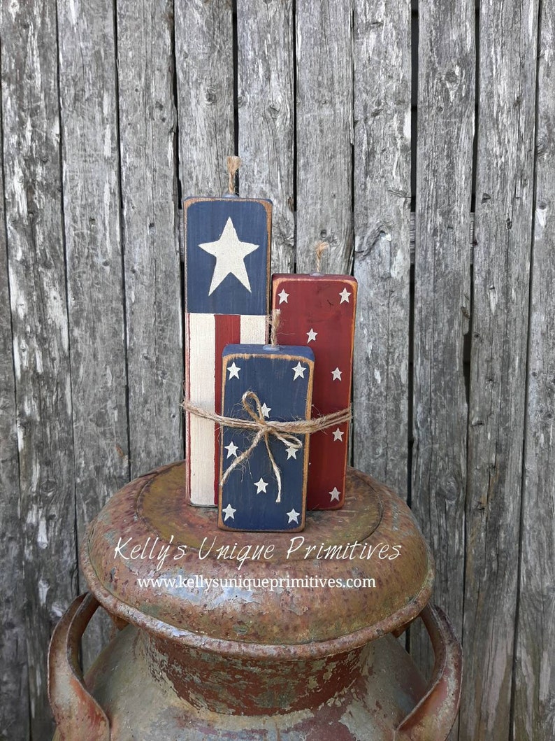 May include: Three wooden blocks painted to look like fireworks with red, white, and blue stars. The blocks are stacked on top of a rusty metal milk can. The text "Kelly's Unique Primitives" and "www.kellyuniqueprimitives.com" is printed on the can.