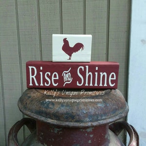 May include: Red wooden sign with the text "Rise and Shine" in white lettering. A smaller white wooden sign with a red rooster silhouette sits on top of the larger sign. The signs are sitting on top of a rusty metal container.