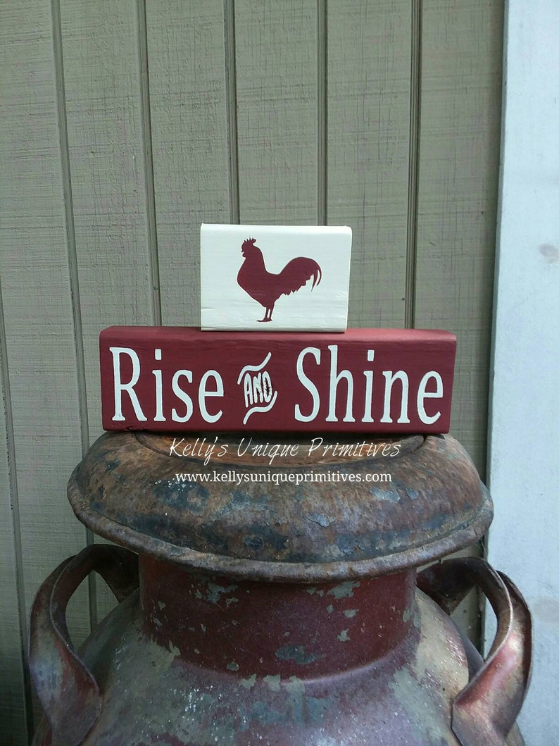 May include: Red wooden sign with the text "Rise and Shine" in white lettering. A smaller white wooden sign with a red rooster silhouette sits on top of the larger sign. The signs are sitting on top of a rusty metal container.