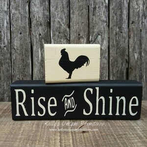 May include: Black wooden block sign with white text that reads "Rise and Shine". A smaller, cream-colored wooden block with a black silhouette of a rooster sits on top of the larger block.