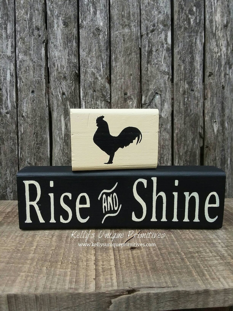 May include: Black wooden block sign with white text that reads "Rise and Shine". A smaller, cream-colored wooden block with a black silhouette of a rooster sits on top of the larger block.