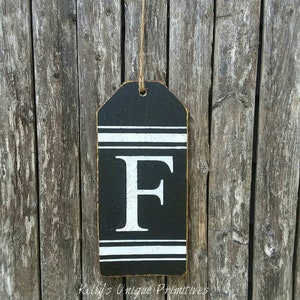 May include: Black wooden sign with a white letter F and white stripes. The sign is hanging from a twine string. The sign says "Kelly's Unique Primitives" and "www.kellysunqiueprimitives.com".