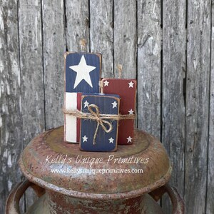 May include: Three wooden blocks painted with red, white, and blue stars and stripes, resembling American flags. The blocks are stacked on top of a rusty metal container. The text "Kelly's Unique Primitives" and "www.kellysuniqueprimitives.com" is printed on the container.