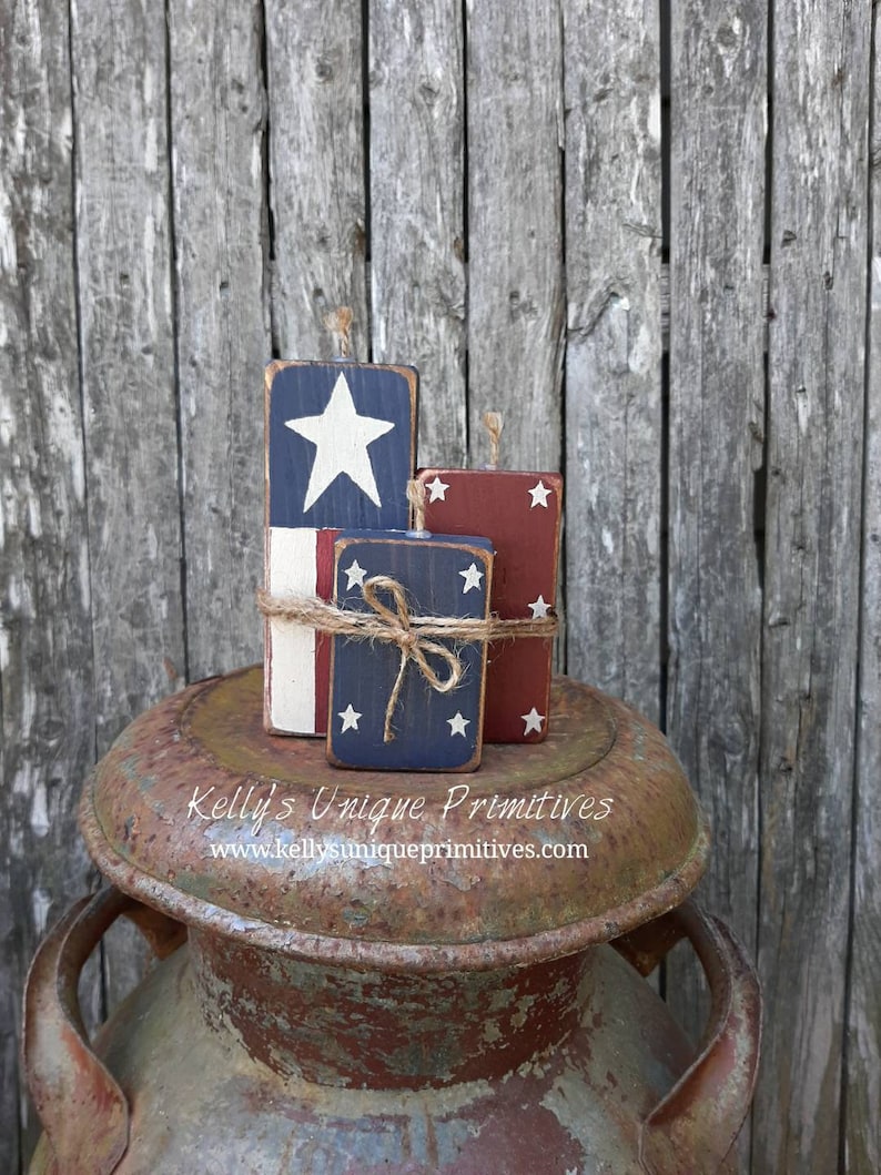 May include: Three wooden blocks painted with red, white, and blue stars and stripes, resembling American flags. The blocks are stacked on top of a rusty metal container. The text "Kelly's Unique Primitives" and "www.kellysuniqueprimitives.com" is printed on the container.